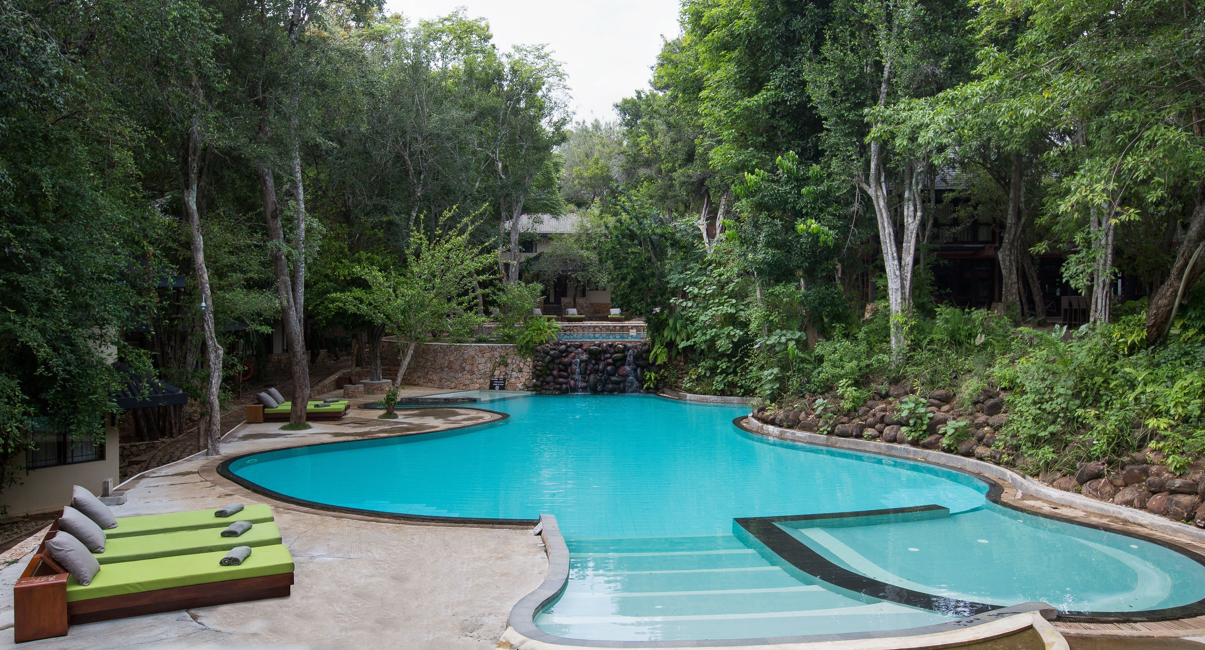 Una piscina de diseño curvo, rodeada de exuberante vegetación y árboles. Se observan sillas de descanso y una pequeña cascada al fondo.