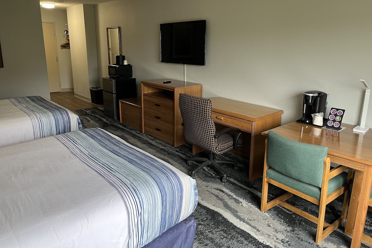 A hotel room with two beds, a desk, a chair, a TV, a coffee maker, and a lamp.