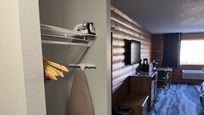 A hotel room with a wooden log wall, a flat-screen TV mounted on the wall, a small desk with a chair, a wooden bench, a trash bin, and a window with curtains. - Days Inn & Suites by Wyndham Williamsburg IA North (Williamsburg)