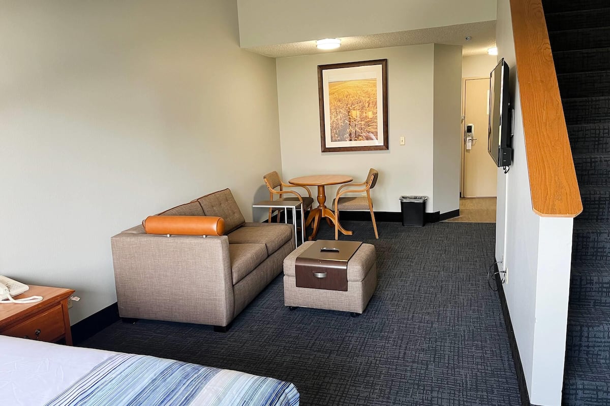 A hotel room with a bed, a sofa, a small table with chairs, a TV mounted on the wall, and a framed picture above the table.