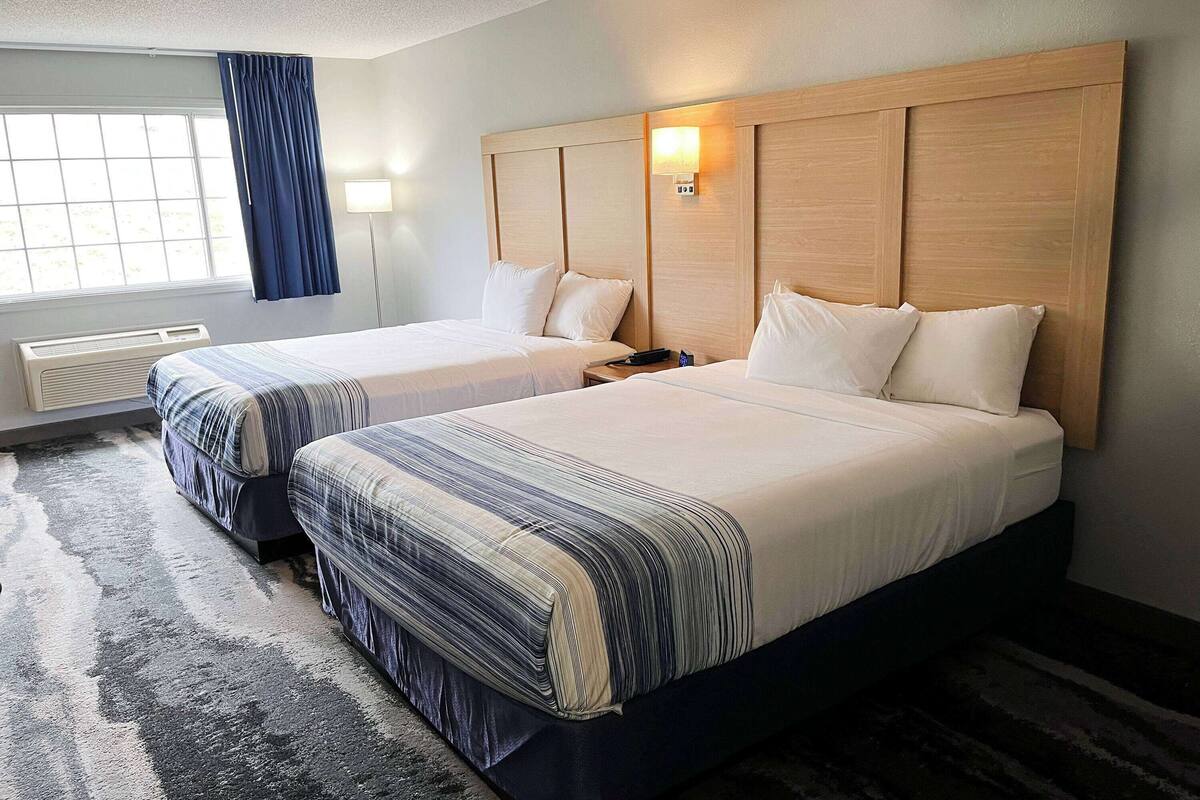 A hotel room with two beds, a large window with curtains, a wall-mounted light, and a rug.