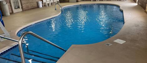 An indoor swimming pool with a curved edge, a metal ladder, and white chairs around the perimeter.