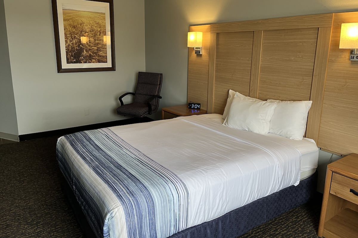 A hotel room with a large bed, a chair, a nightstand, a wall clock, and a framed picture.