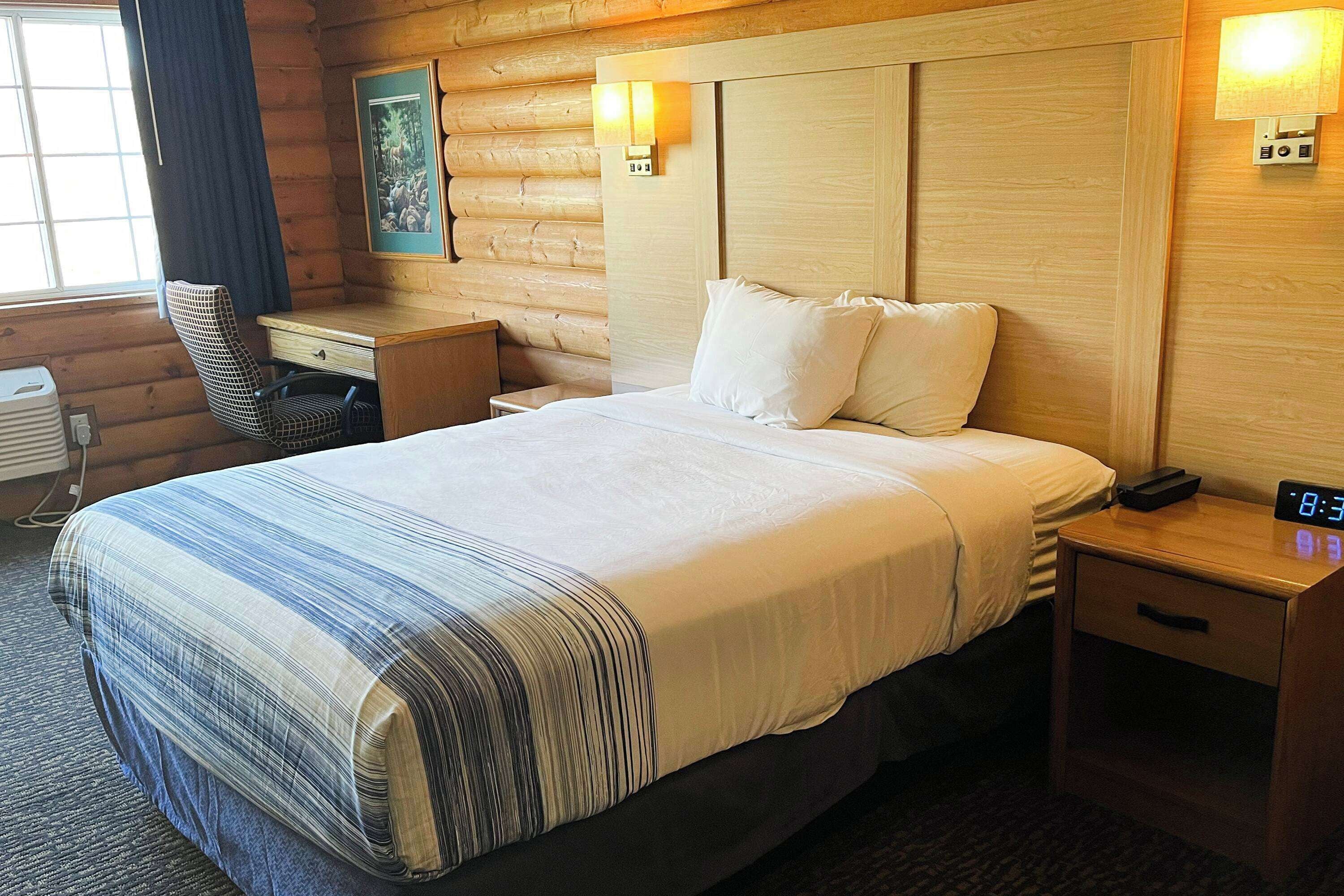 Una cama bien hecha con colcha a rayas, cabecera de madera, lámparas de noche, mesita de noche, una silla e imagen en un marco en la pared.