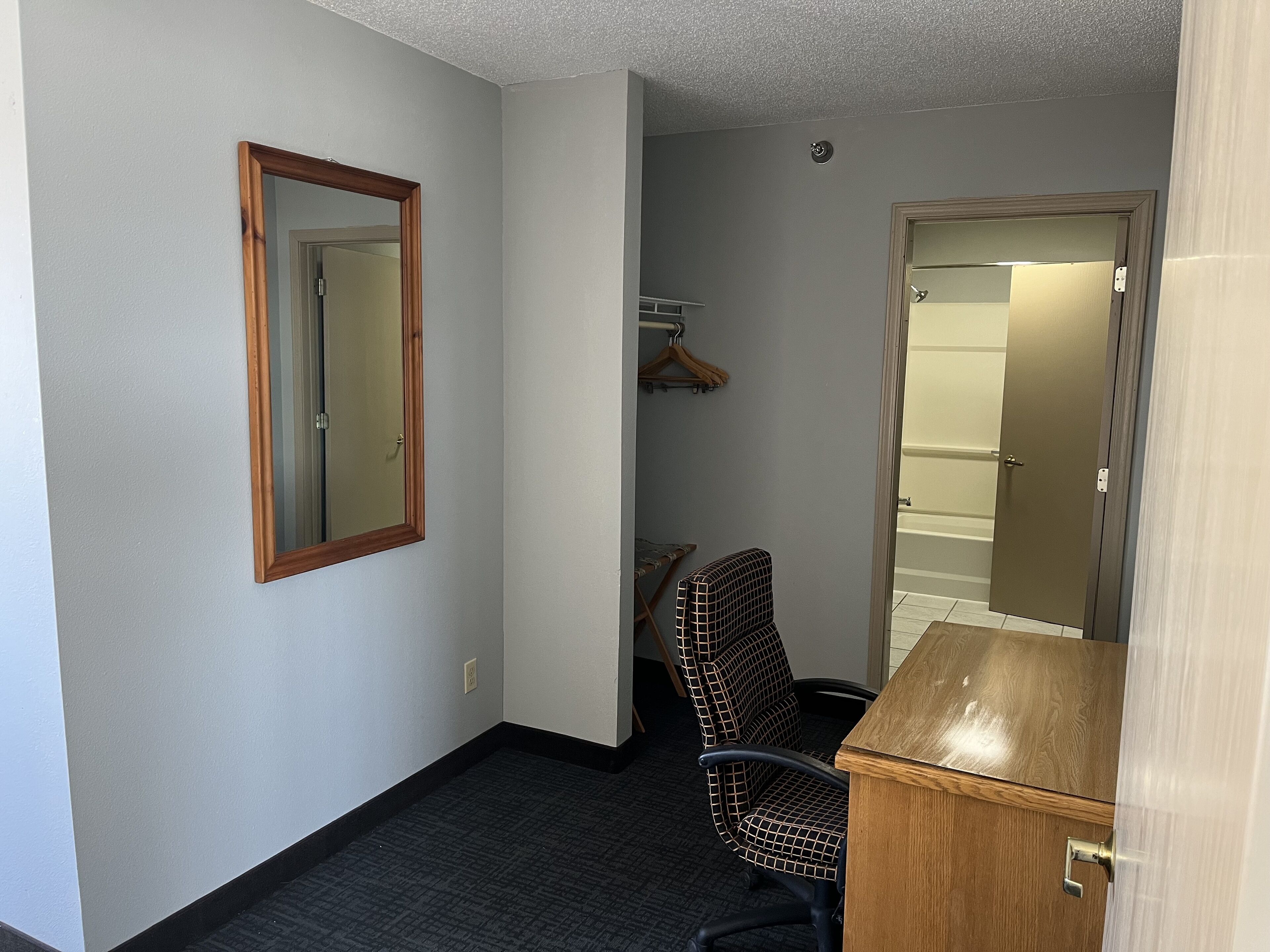 Habitación de hotel con un escritorio de madera, una silla, un espejo y una puerta que da a un baño.