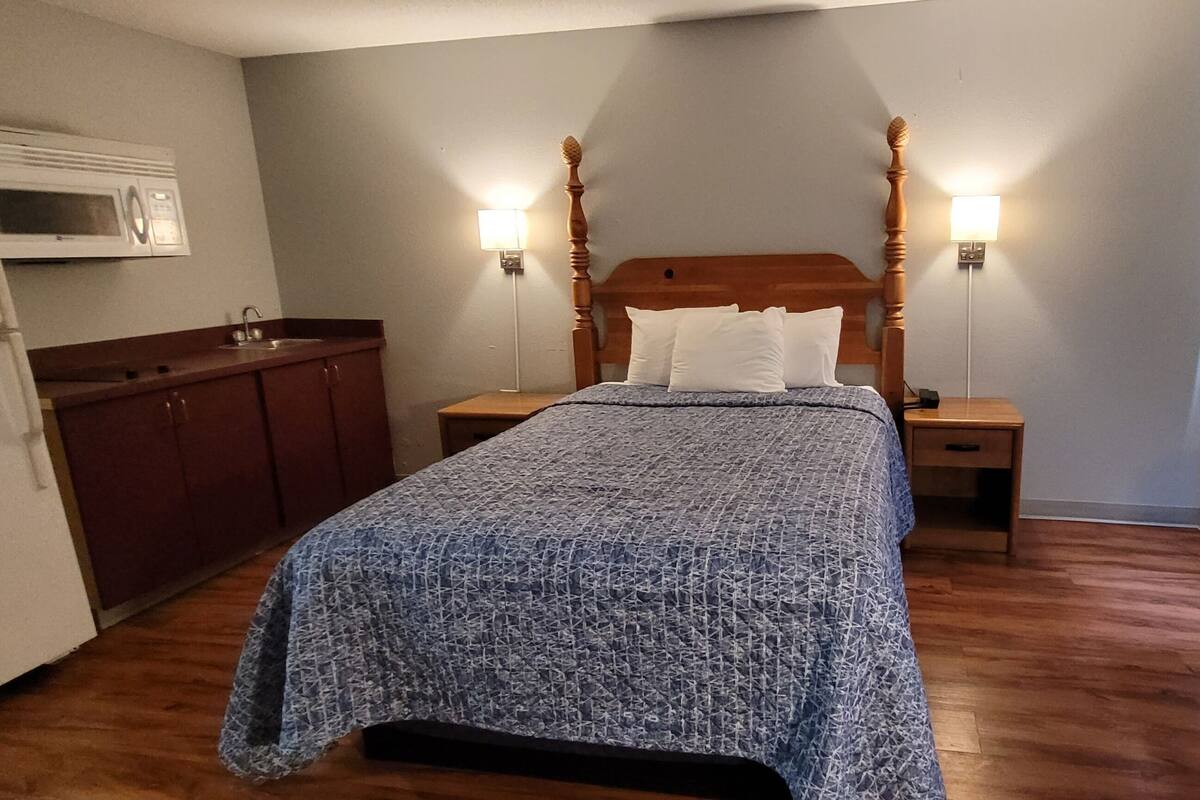 A compact hotel room with a bed, a nightstand, a wooden headboard, a microwave, a refrigerator, a window with curtains, and a heater.