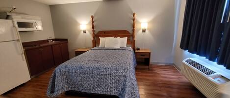 A compact hotel room with a bed, a nightstand, a wooden headboard, a microwave, a refrigerator, a window with curtains, and a heater.