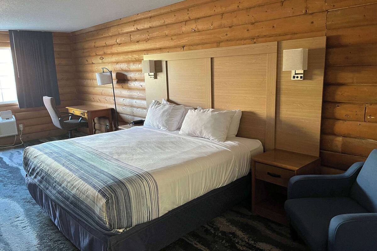 A hotel room with a wooden log wall, a large bed with white linens, a desk with a chair, and a small table with a lamp.