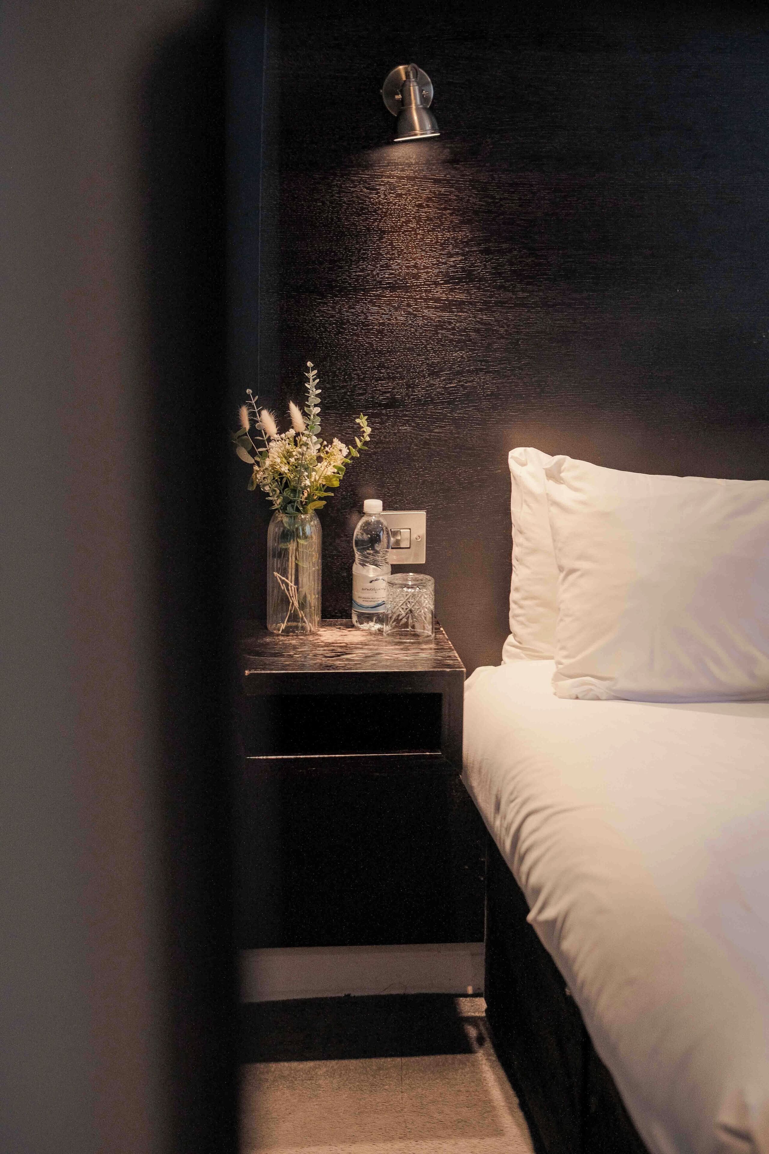 A bedroom with a bed, a nightstand, a lamp, a vase with flowers, and a bottle.