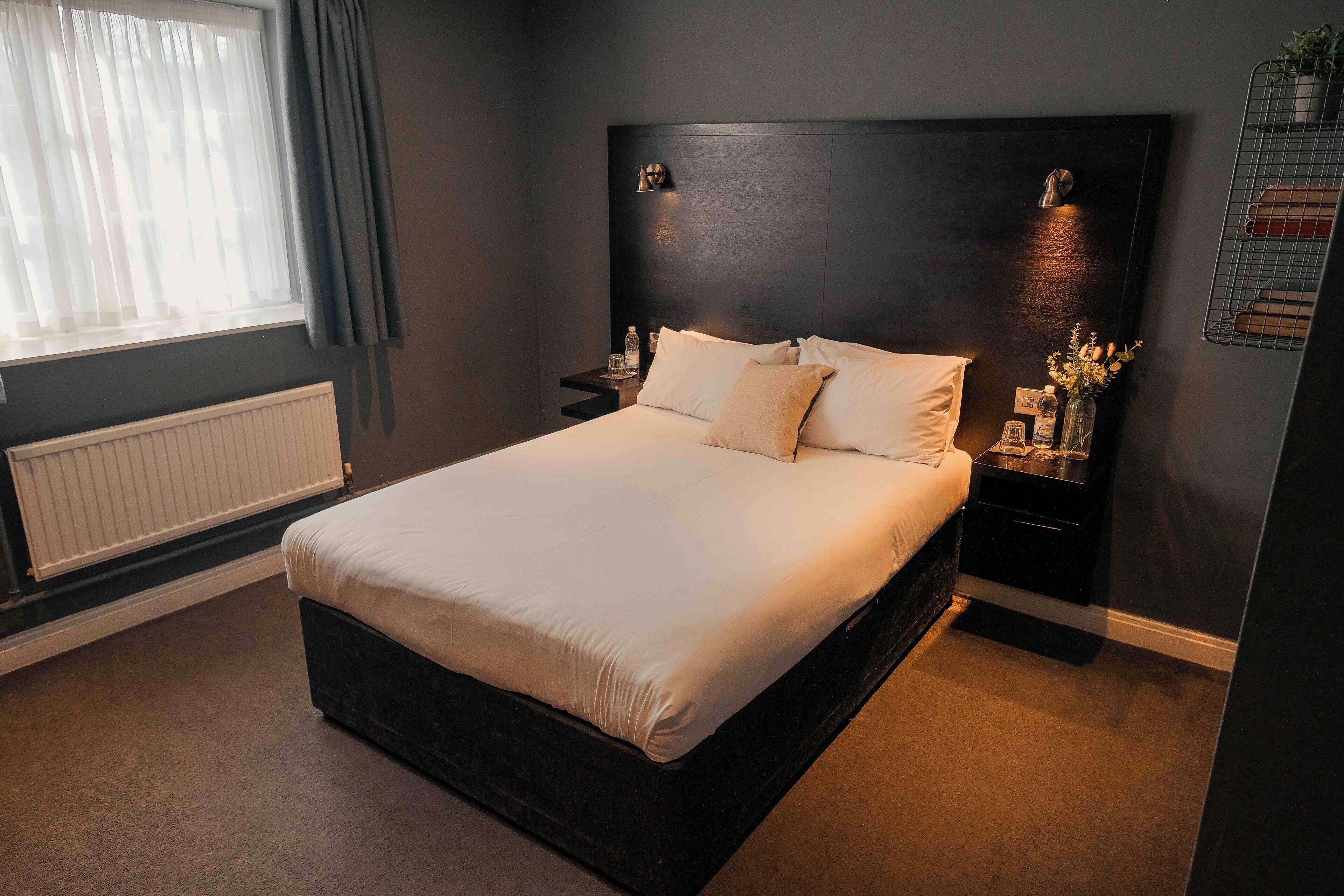 A neatly made bed in a room with a dark headboard, a bedside table with a lamp, and a window with curtains.