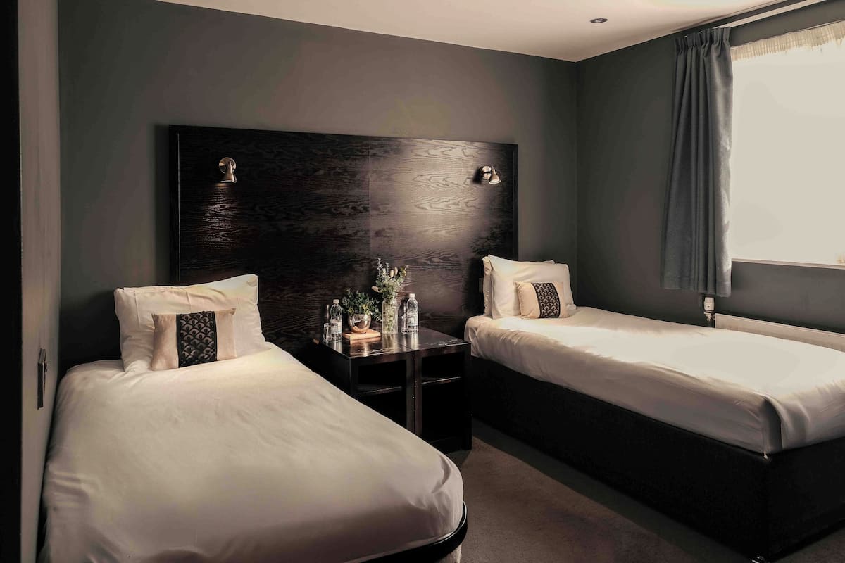 A hotel room with two single beds, a dark wooden headboard, bedside tables with lamps, and a window with curtains.