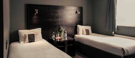 A hotel room with two single beds, a dark wooden headboard, bedside tables with lamps, and a window with curtains.