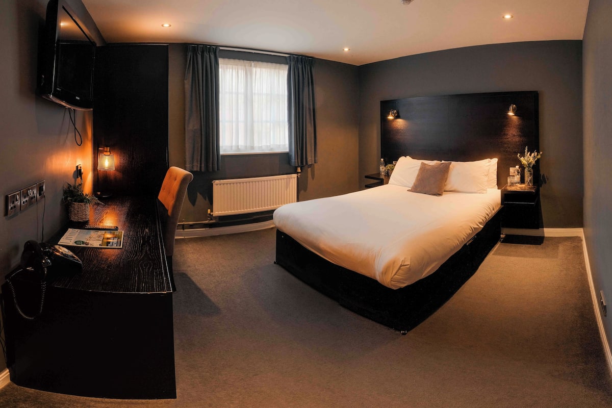A modern hotel room with a large bed, a desk with a chair, a TV mounted on the wall, and a window with curtains.