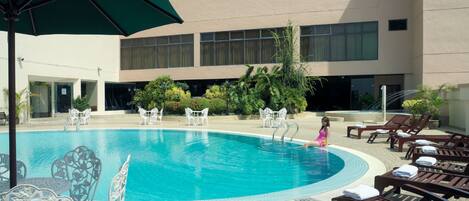 Una piscina con sillones y mesas, rodeada por un edificio con amplios ventanales y un jardín ajardinado.
