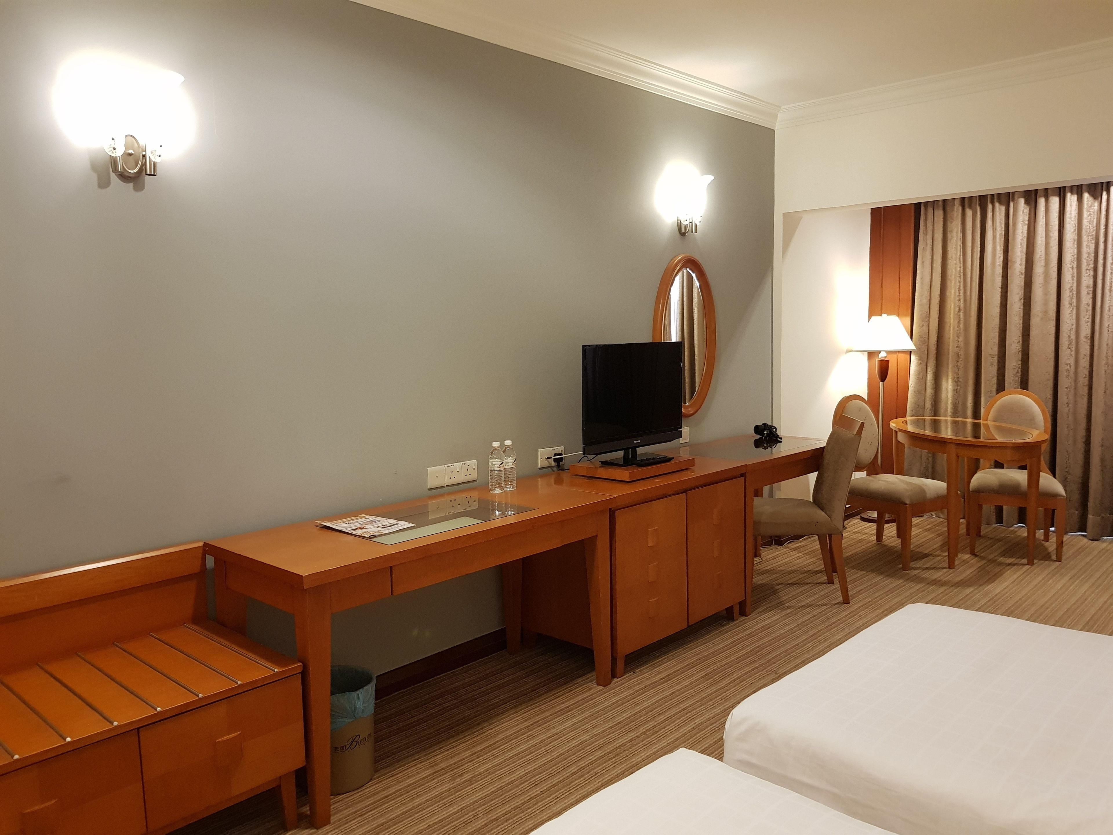 Habitación de hotel con escritorio de madera, televisor, silla, mesita de noche con lámpara y dos camas.