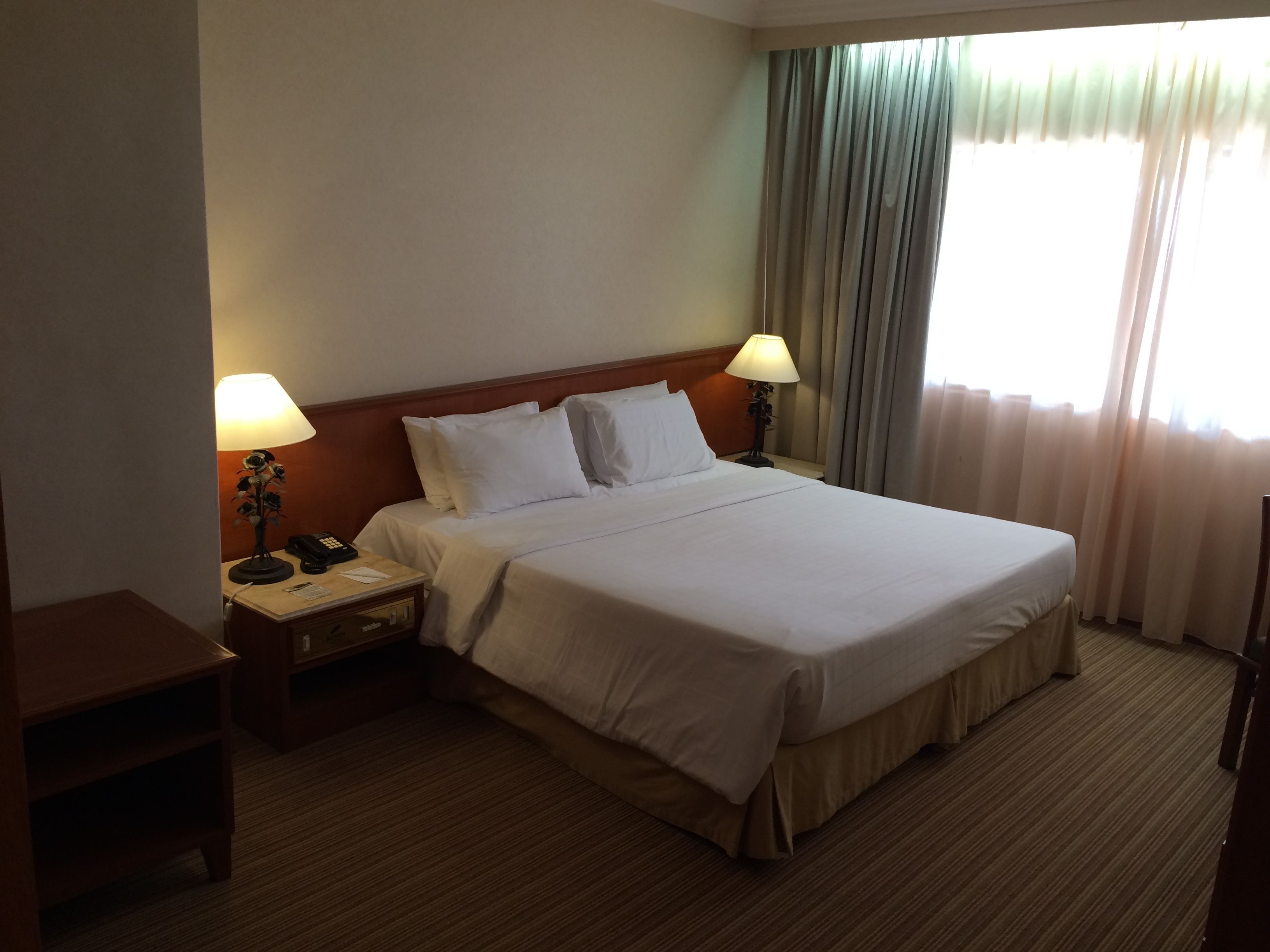 A hotel room with a large bed, two bedside tables with lamps, a wooden headboard, and a window with curtains.
