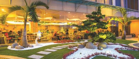 An indoor dining area with tables and chairs, a decorative tree, and a well-maintained garden with rocks and plants.