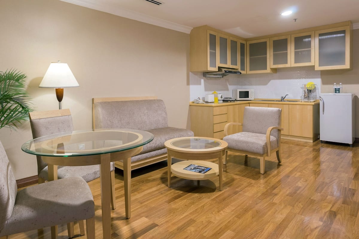 A compact living space with a sofa, two armchairs, a glass-top coffee table, a lamp, a potted plant, a microwave, a refrigerator, and wooden cabinets.