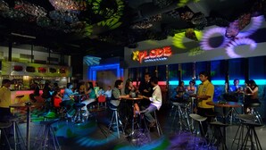 En bardel med barstolar och bord, en neonreklamskylt med texten ”Xplode Dance Club” och ett tak med hängande dekorationer.