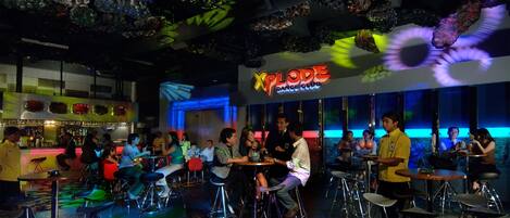 A bar area with stools and tables, a neon sign reading "Xplode Dance Club", and a ceiling with hanging decorations.