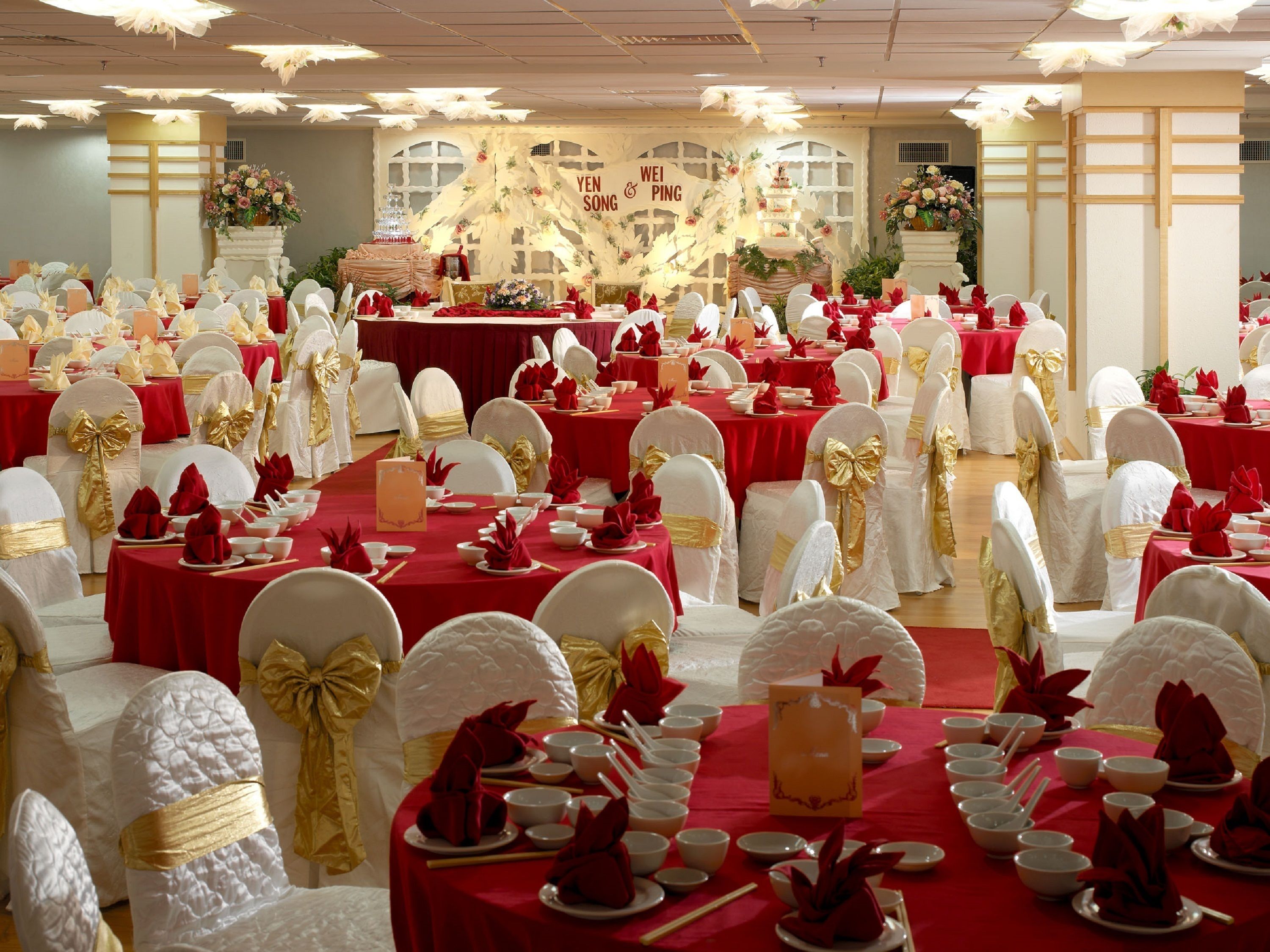 Een balzaal met gedekte tafels voor een formeel evenement, met rode tafelkleden en witte stoelen met gouden linten.