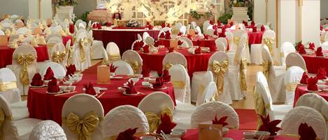 Salão de banquetes com mesas montadas para um evento formal, com toalhas vermelhas e cadeiras brancas adornadas com fitas douradas.