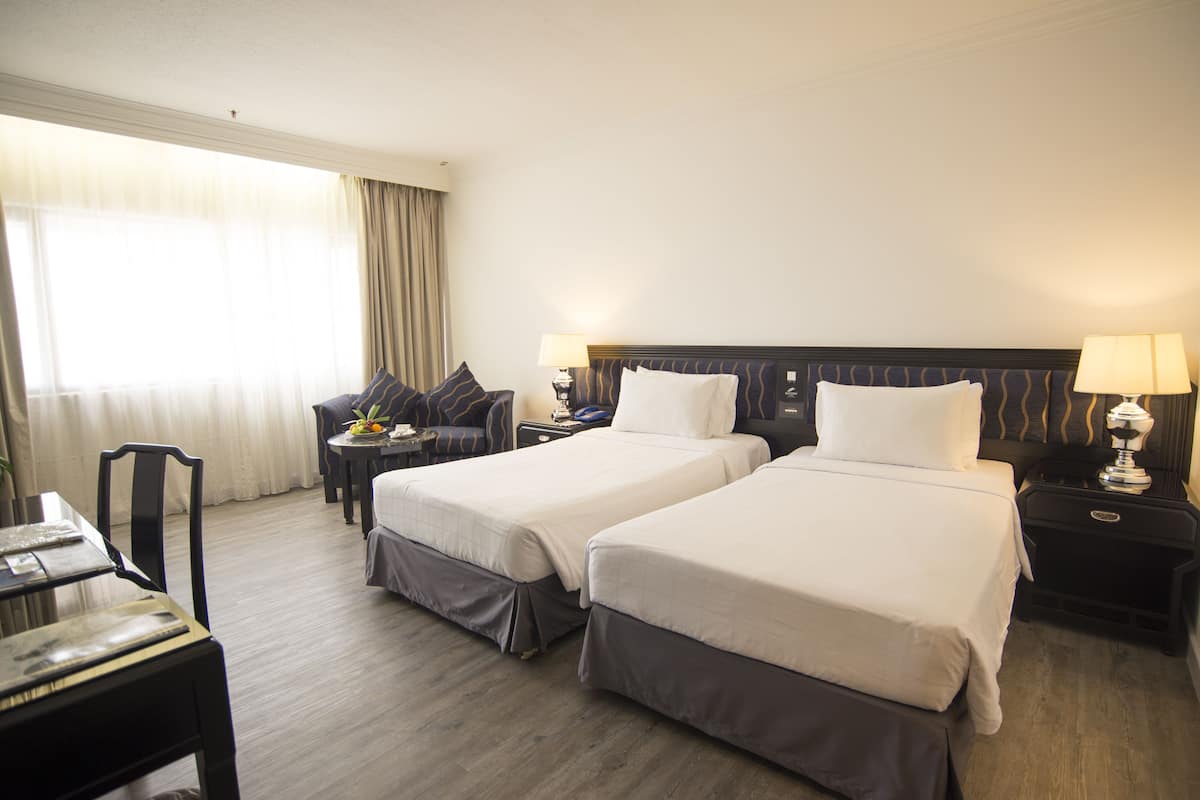 A hotel room with two beds, a desk, a chair, a nightstand with a lamp, and a window with curtains.