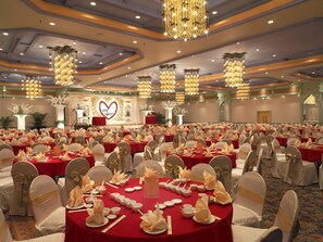 Uma sala de banquetes com mesas redondas dispostas para um evento formal, apresentando um palco com um painel em formato de coração e decoração floral.