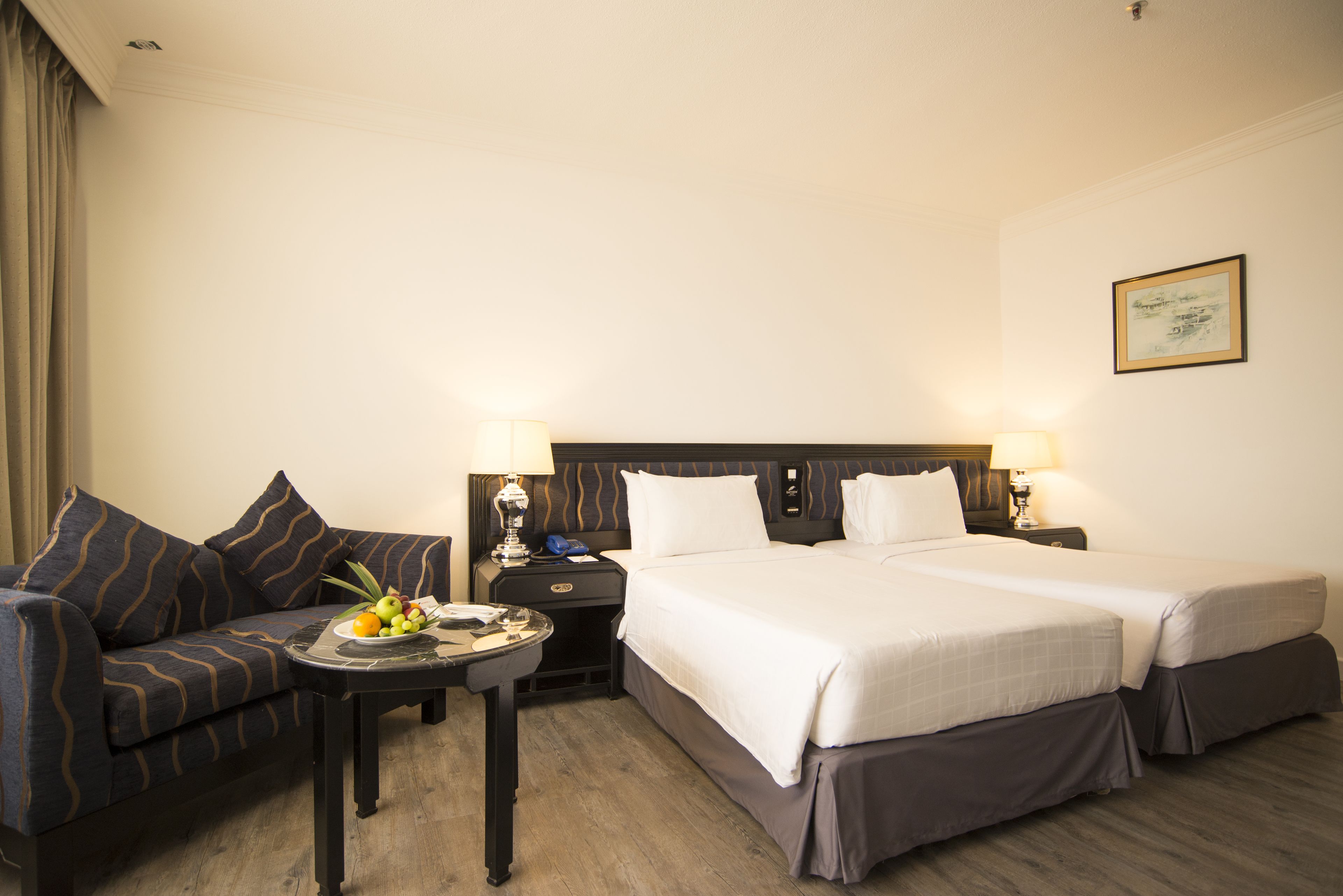 A hotel room with a double bed, a sofa, a small table with fruit, two bedside lamps, and a framed picture on the wall.