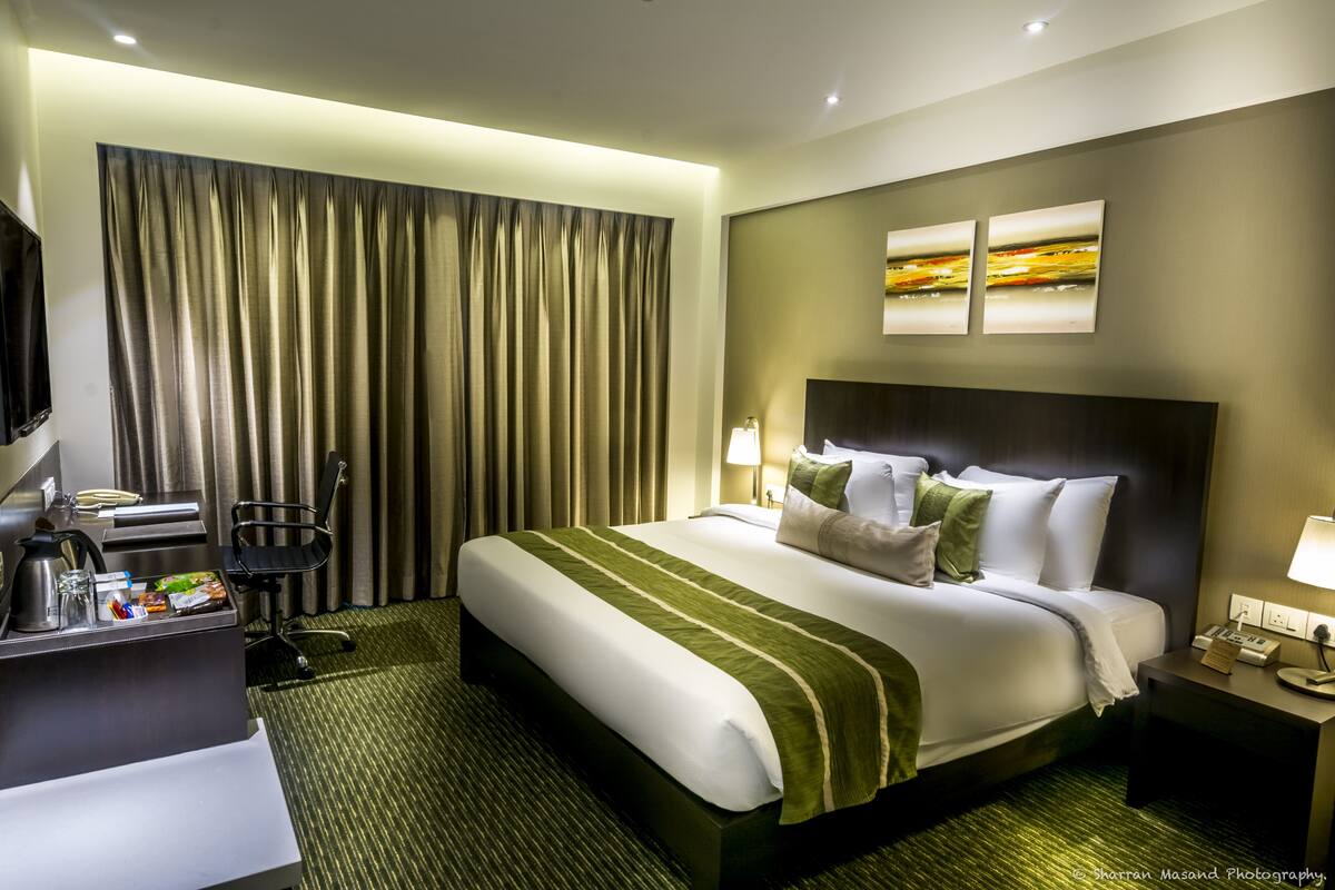 A hotel room with a large bed, a desk with a coffee maker, a chair, and two paintings on the wall.