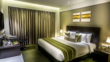 Quarto de hotel com uma cama grande, uma escrivaninha com cafeteira, uma cadeira e duas pinturas na parede.
