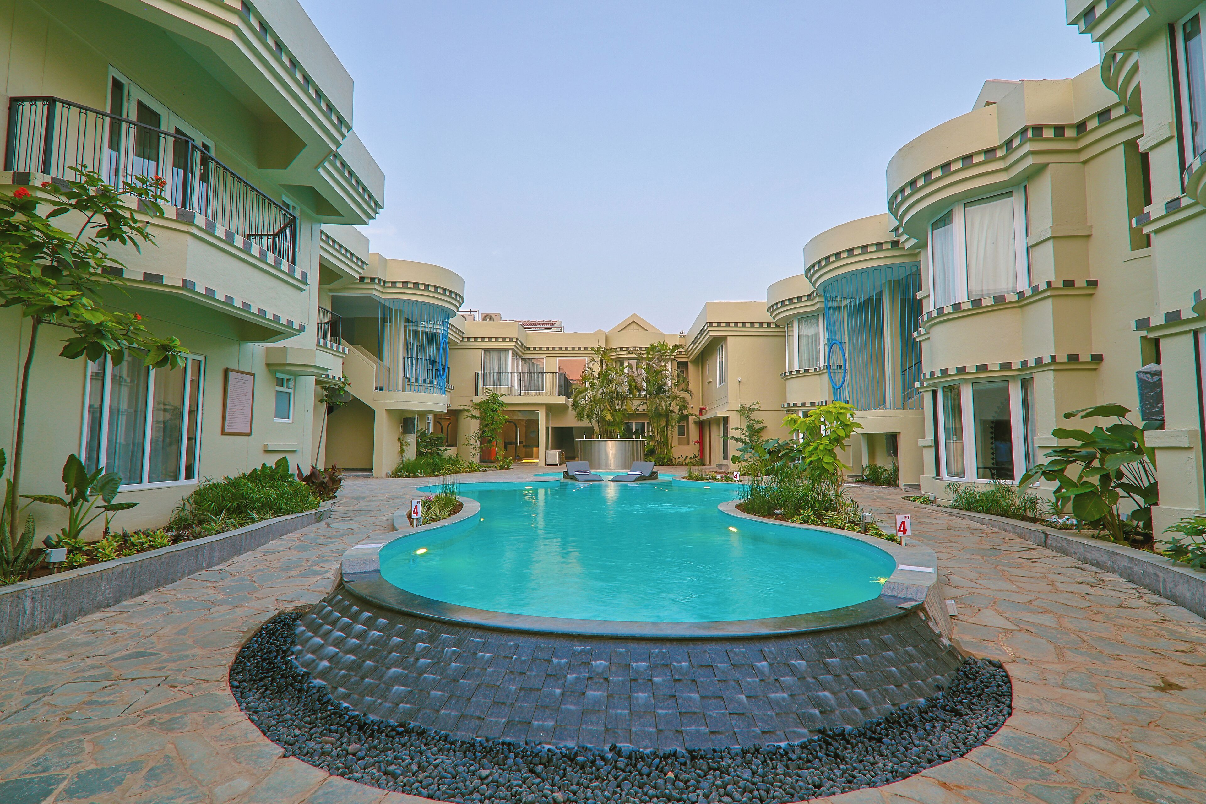 A courtyard with a swimming pool surrounded by a stone walkway, a fountain, and buildings with balconies.