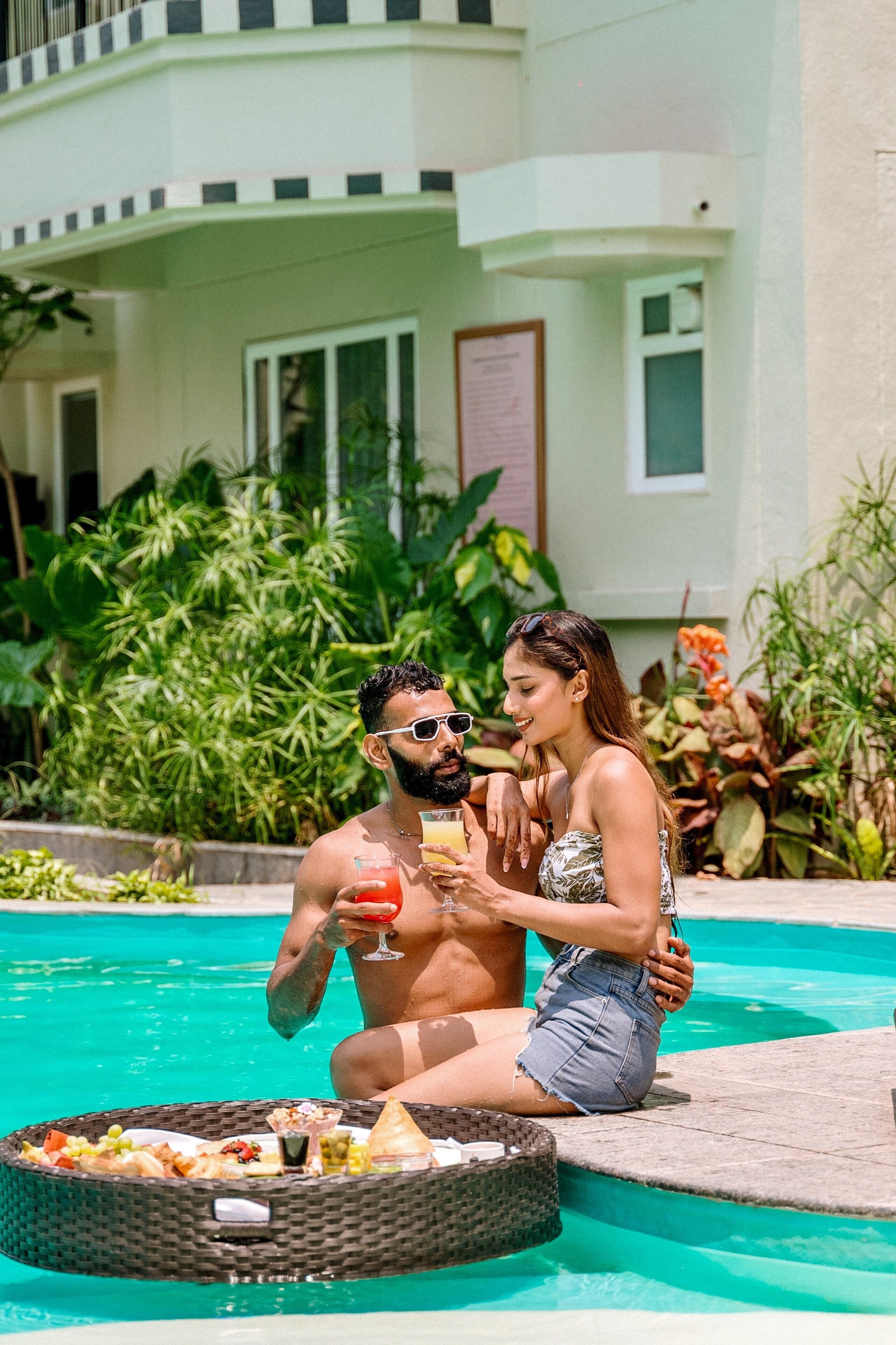 A couple sitting by a pool with drinks and a basket of food.