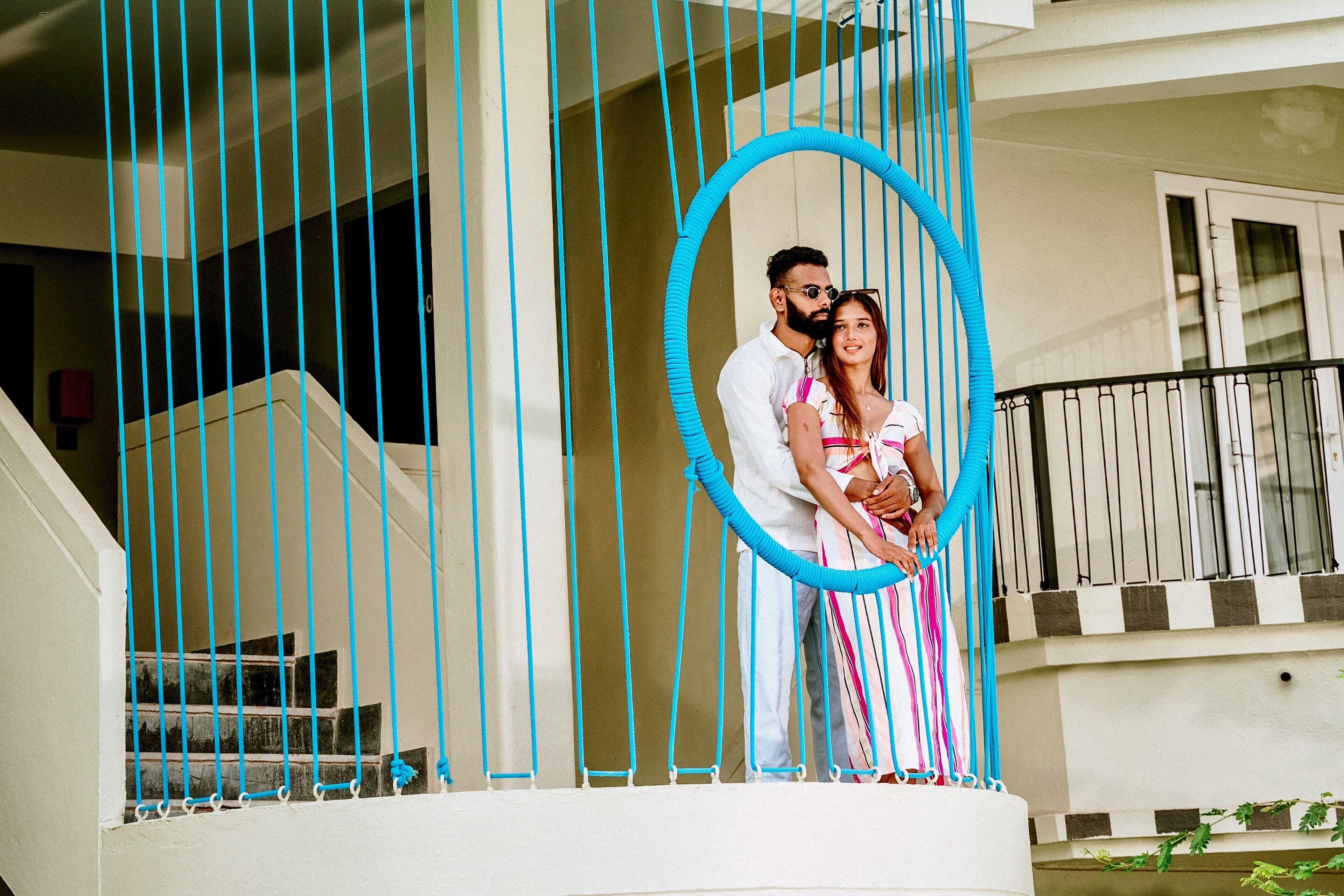 Un couple se tient sur un balcon orné d’un cadre circulaire bleu, avec un escalier et une balustrade.