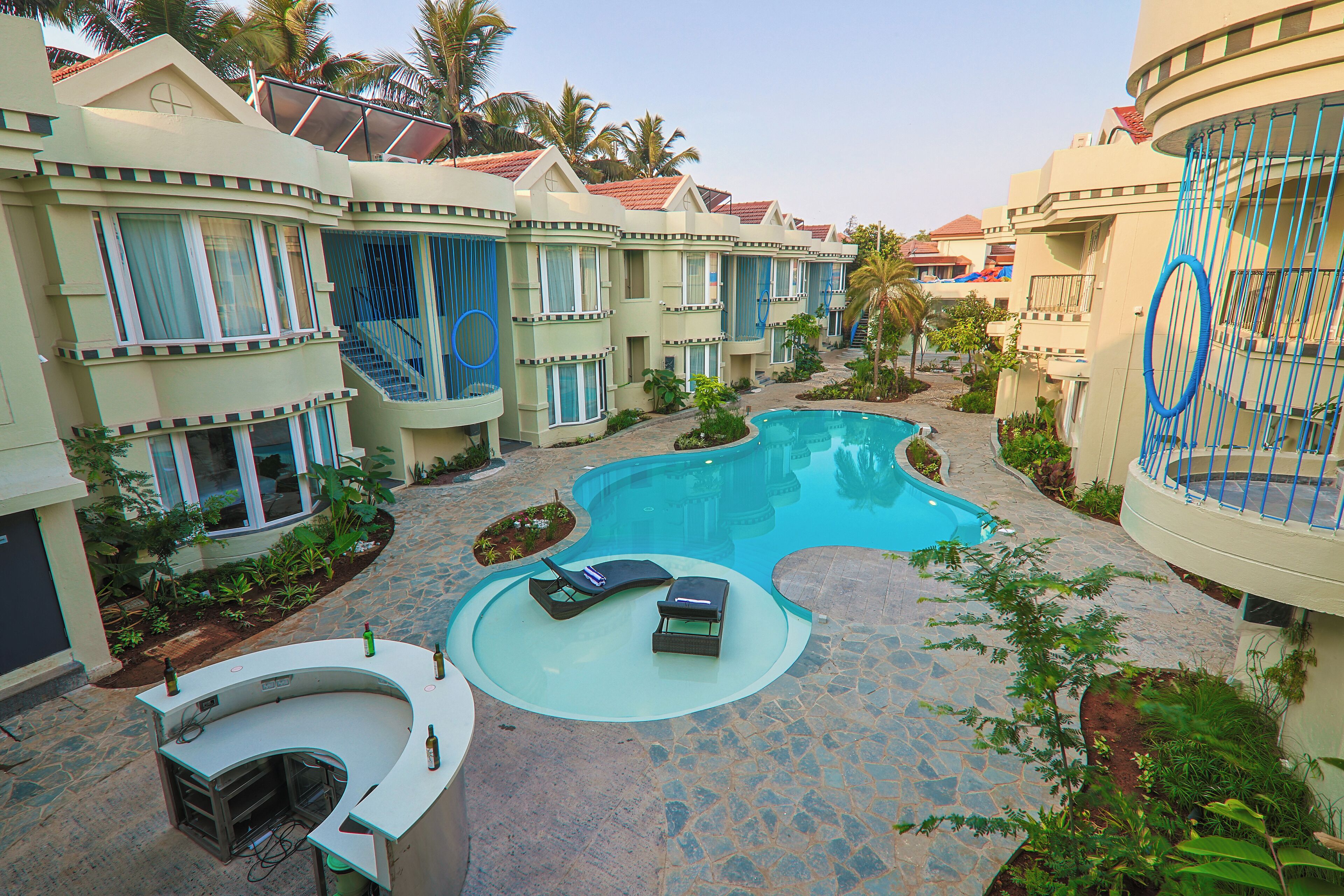 A pool area with lounge chairs and a bar, surrounded by resort-style buildings with balconies and a clear blue sky.