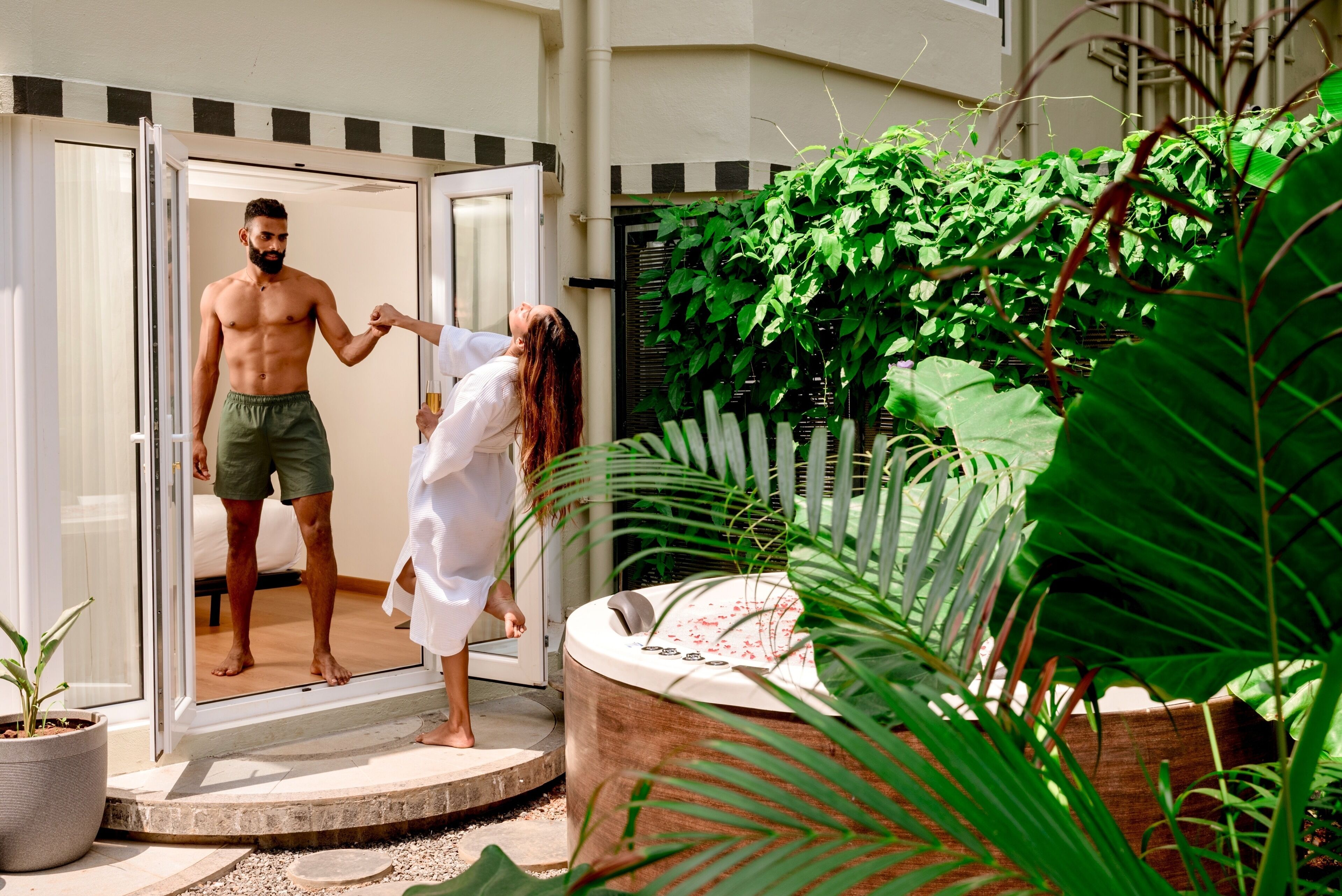 A man and a woman at a hotel entrance, with a hot tub and lush greenery in the background.