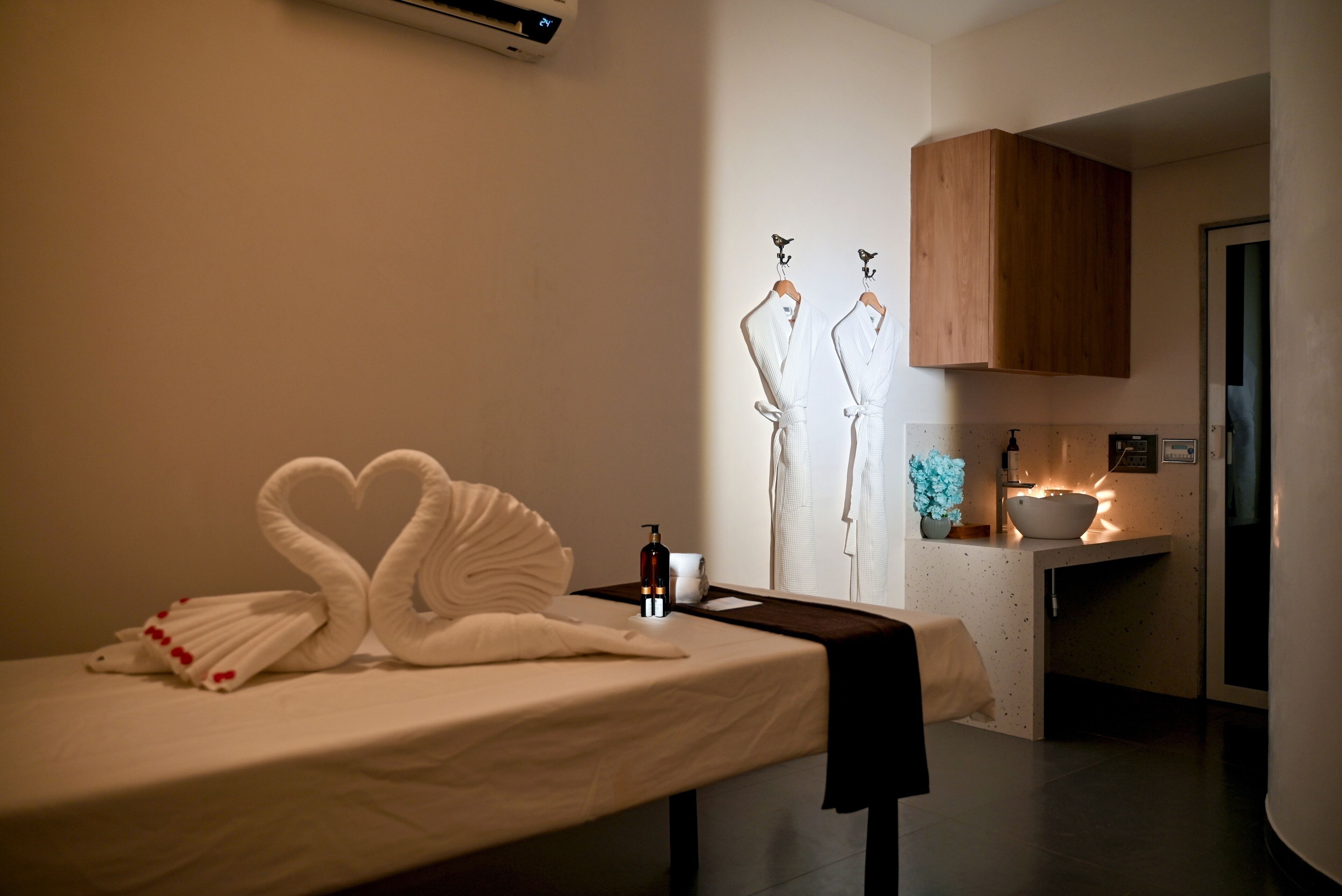 Une chambre d’hôtel comprenant un lit, un porte-serviettes avec des peignoirs, une petite table avec une bouteille et une tasse, et un meuble en bois.
