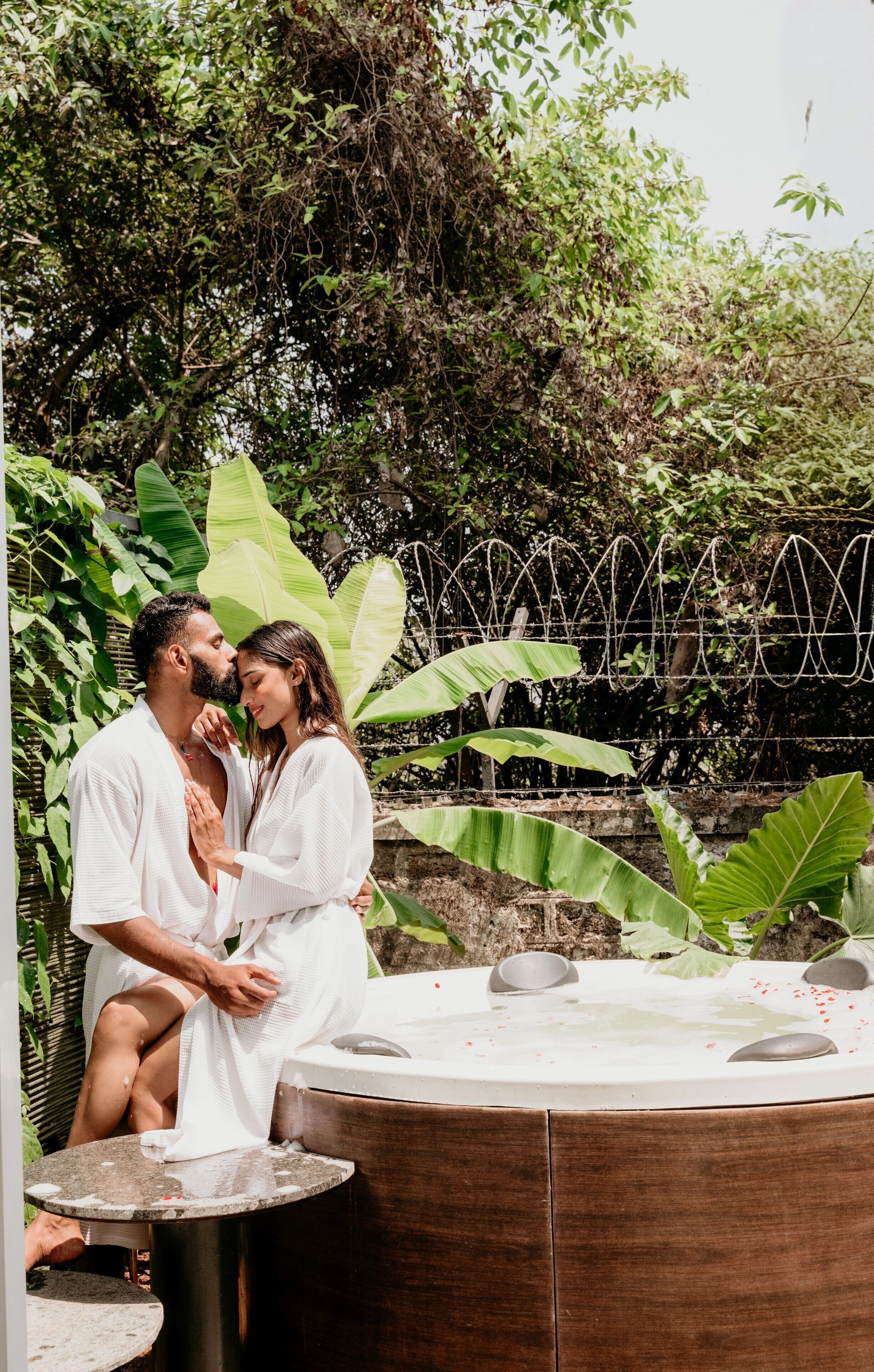 Una pareja sentada junto a un jacuzzi en un jardín exuberante con una terraza de madera y una valla de metal.