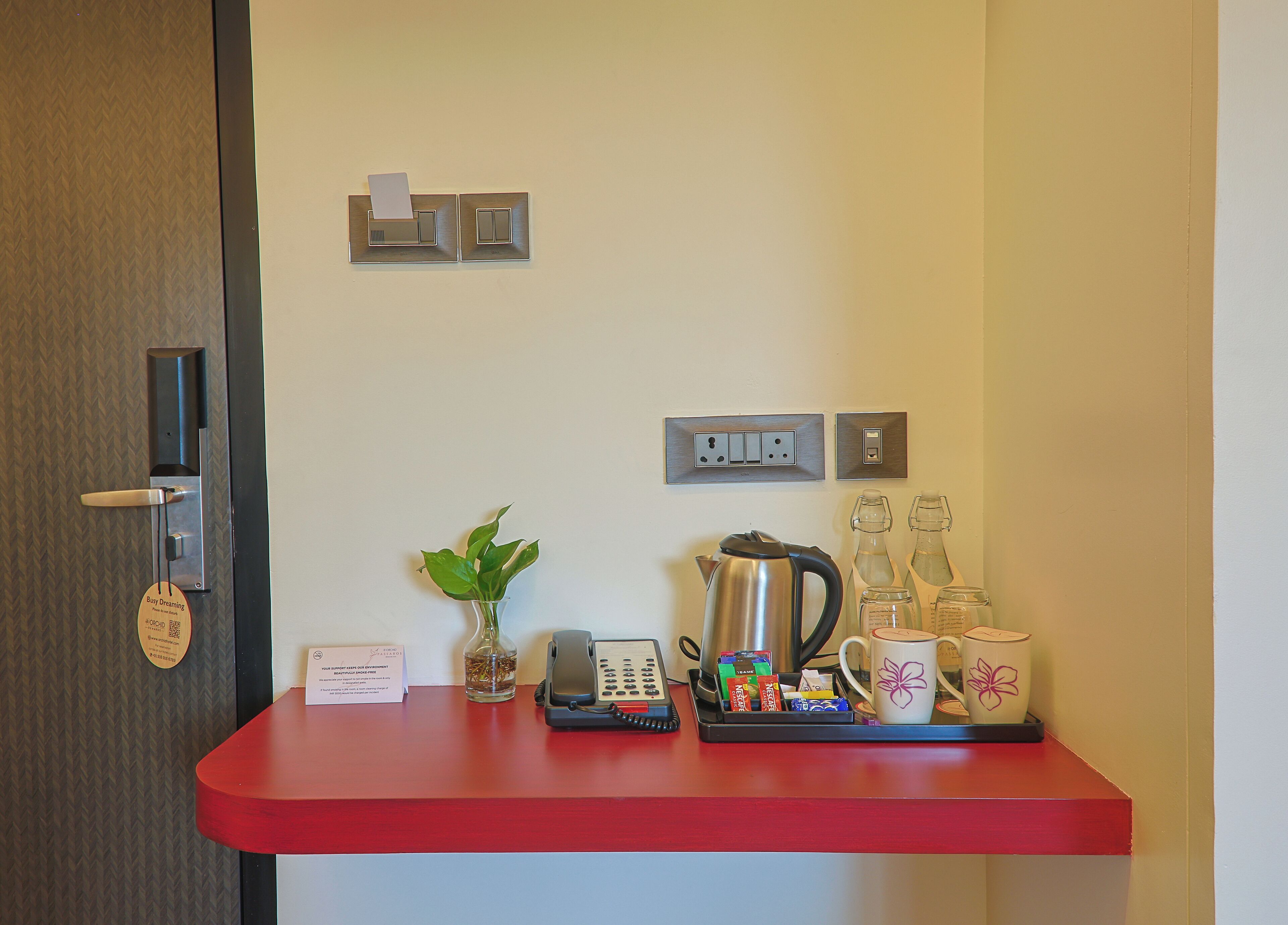 A hotel reception desk with a red counter, a plant, a kettle, a phone, and various coffee mugs and containers.