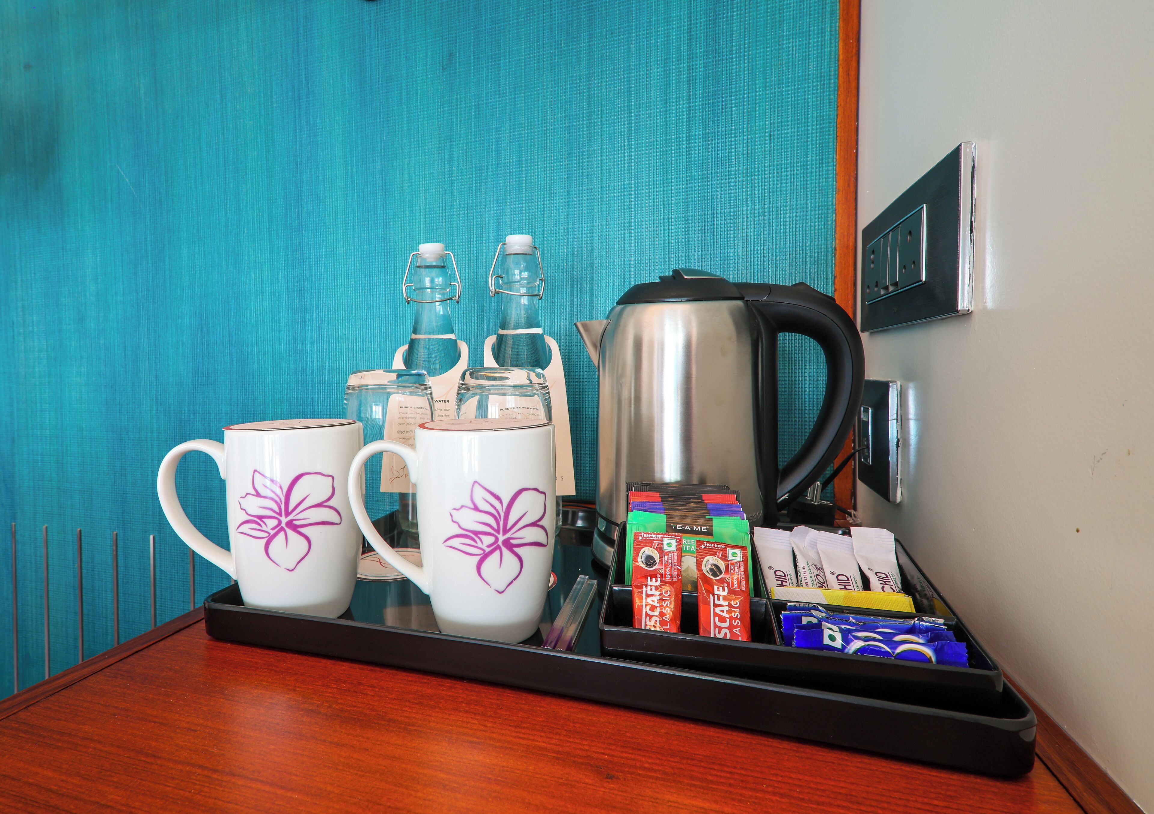Un plateau contenant des tasses à café, des bouteilles d’eau, une bouilloire et des sachets de thé, posé sur une table en bois.
