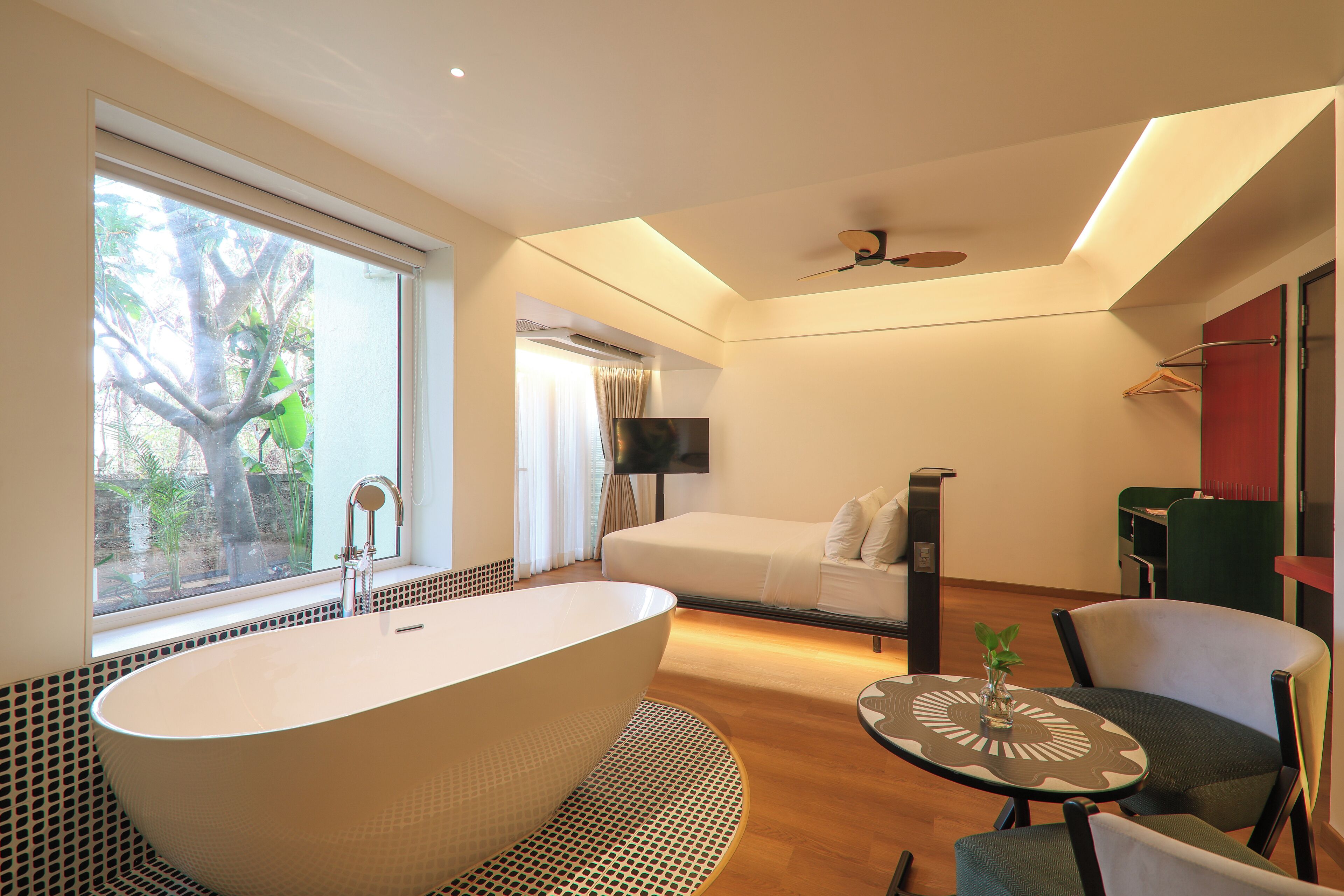 A modern bathroom with a freestanding bathtub, a window with a view of greenery, a bed with a TV, and a small table with a plant.