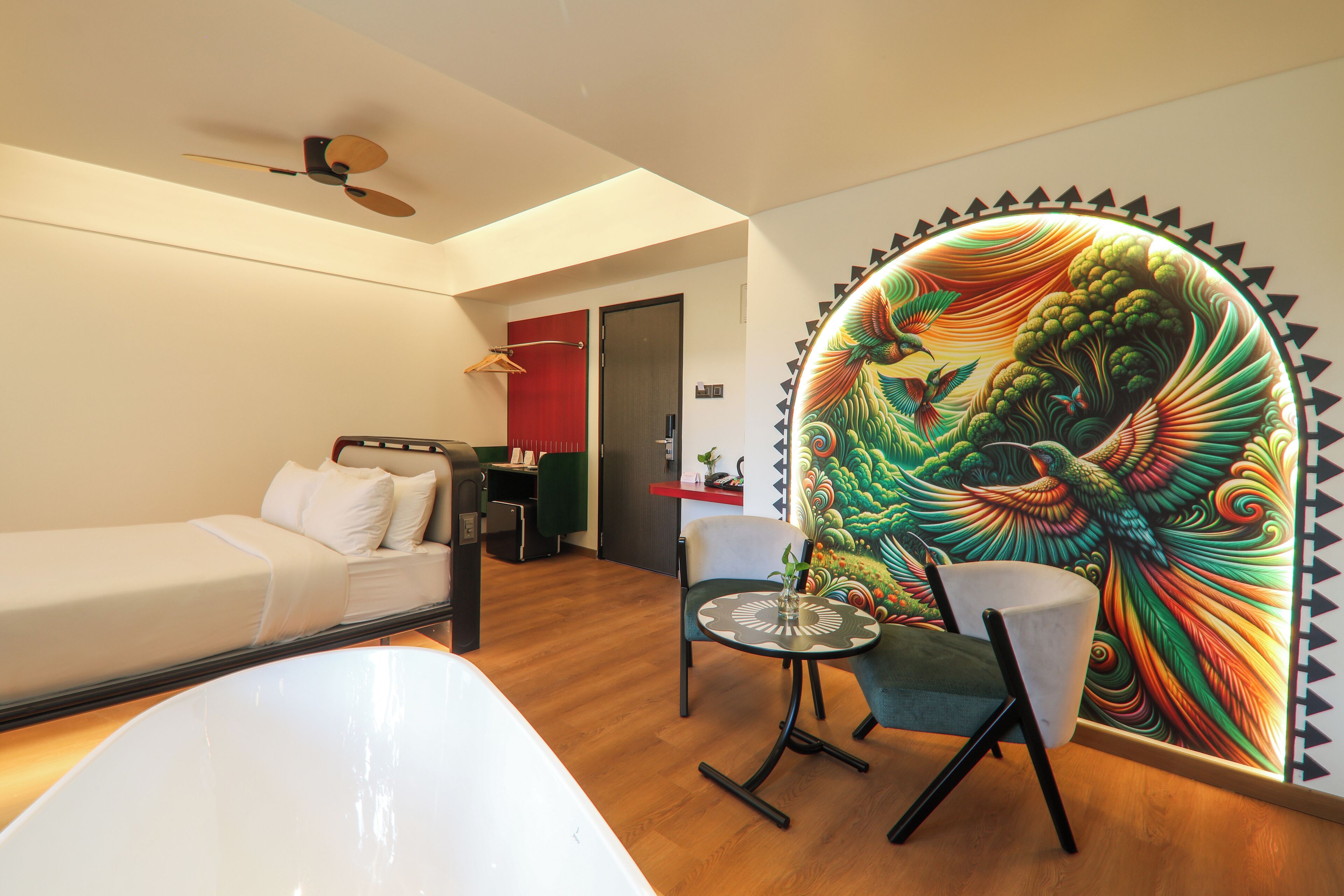 A modern hotel room with a bed, a desk, a chair, and a colorful mural on the wall.