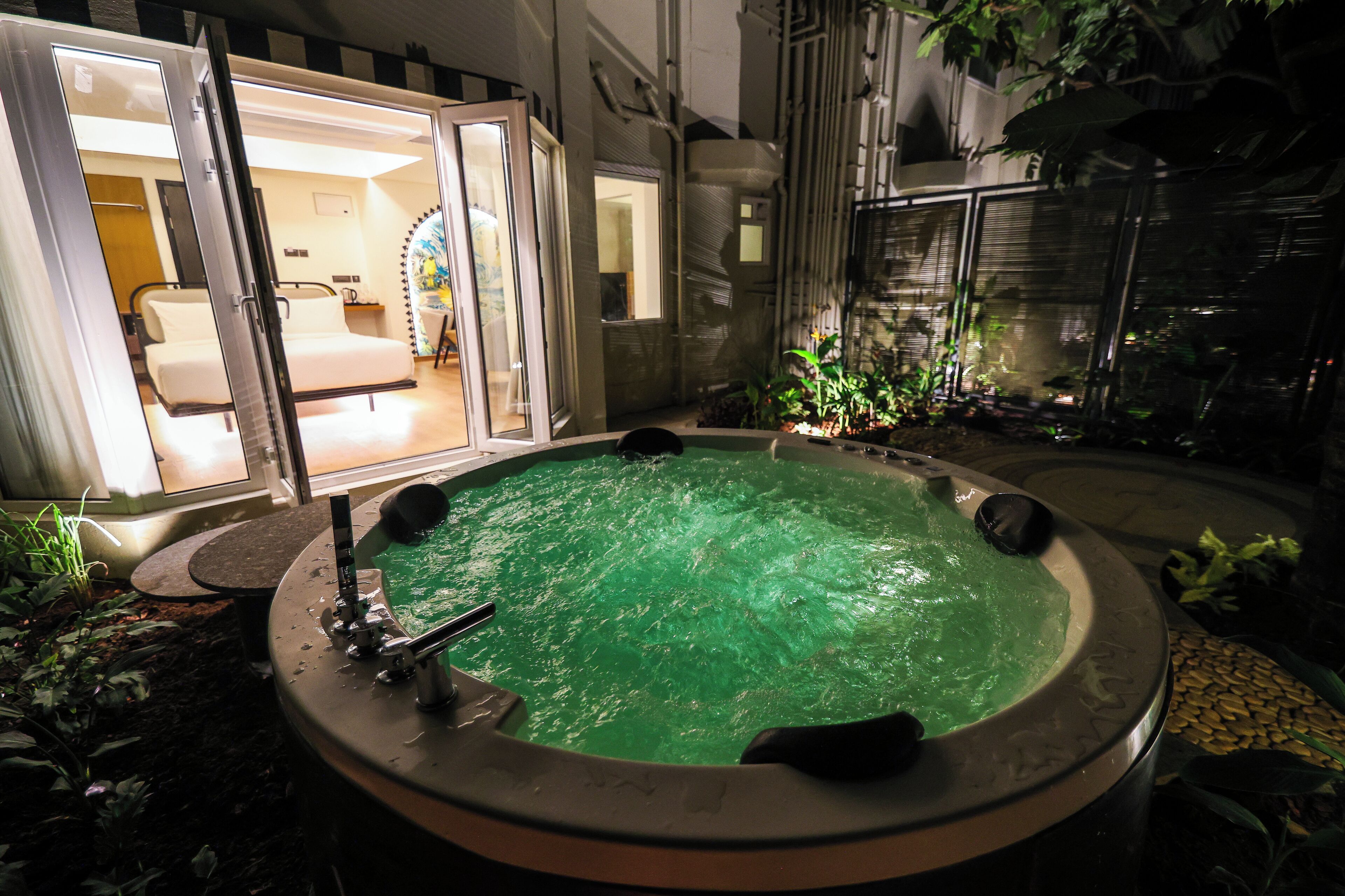 Un bain à remous avec un élément décoratif aquatique à l’eau verdâtre, entouré de plantes et agrémenté d’un petit espace salon.