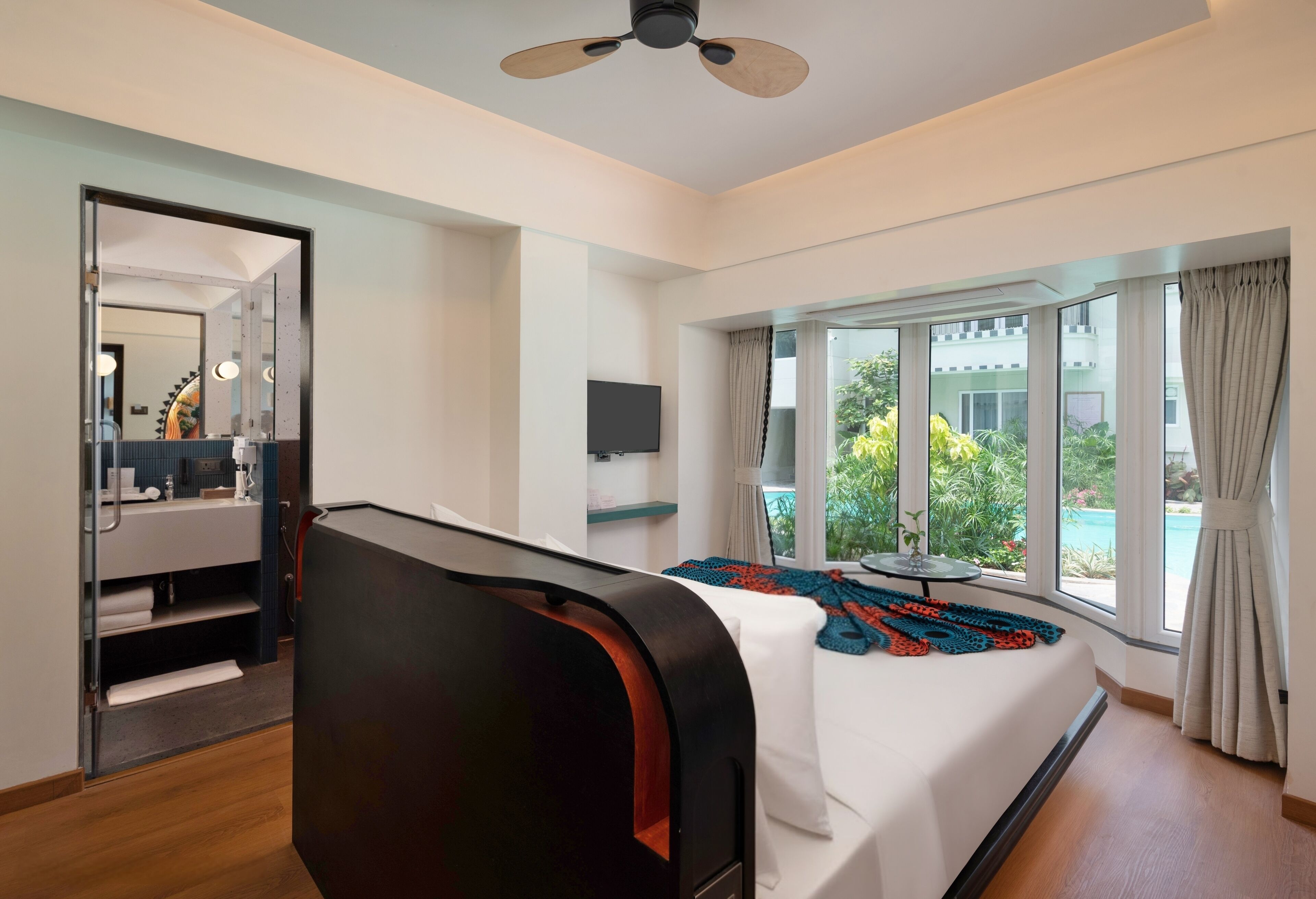 A hotel room with a bed, a ceiling fan, a TV, a window with a view of greenery and a building, and a bathroom with a mirror and towel rack.