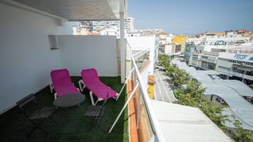 A balcony with two lounge chairs and a small table, overlooking a cityscape with buildings and a clear sky.