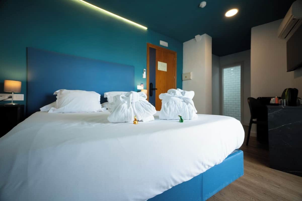 A hotel room with a large bed, a desk with a chair, a door, and a small table with a lamp.
