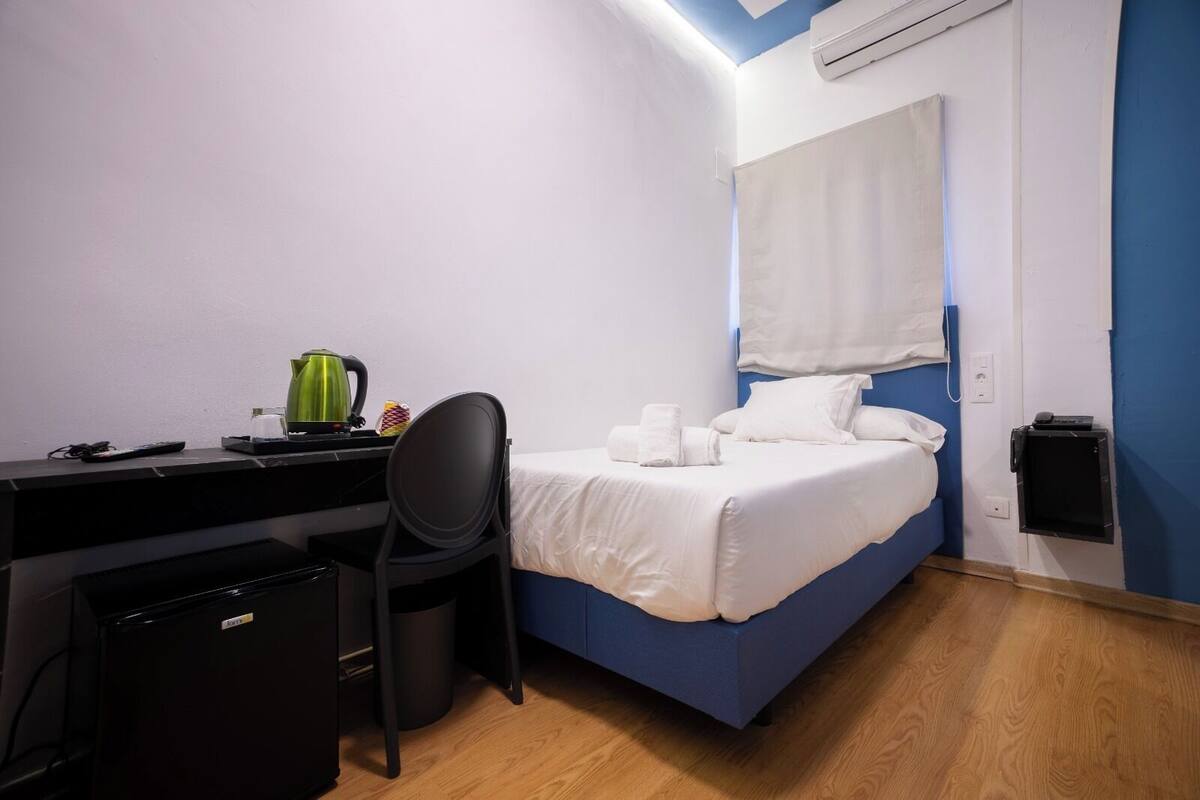 A hotel room with a bed, a desk, a chair, a trash bin, and an air conditioning unit.