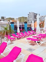 A rooftop pool area with pink lounge chairs and umbrellas, a palm tree, and a building labeled 'HOTEL SIREN.'