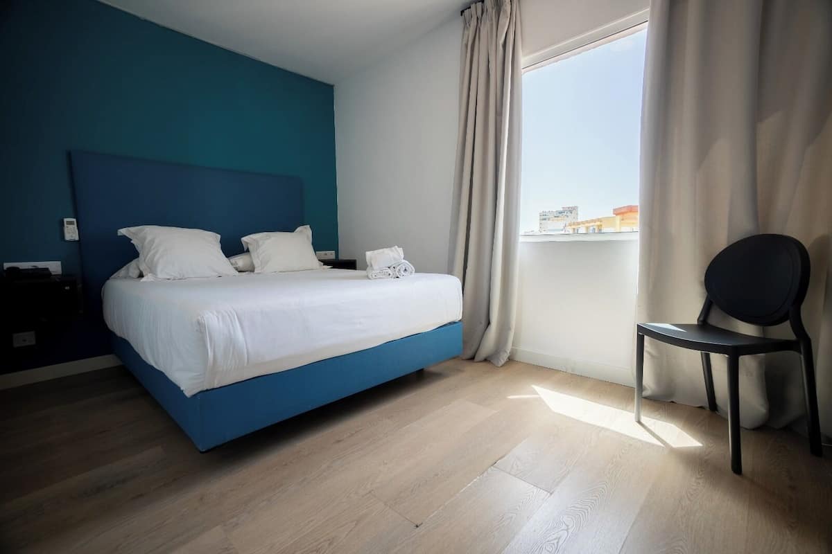 A modern hotel room with a large bed, a chair, a nightstand, and a view of the city through the window.
