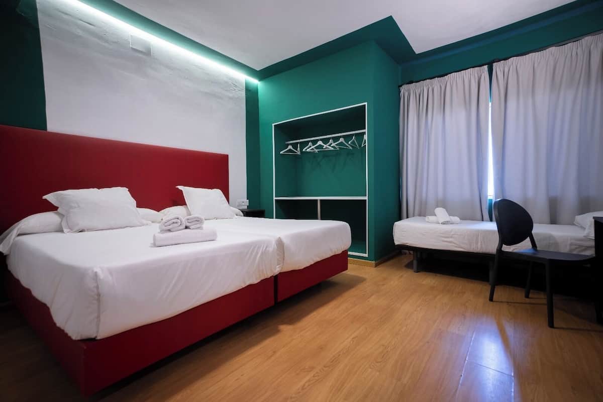 A hotel room with a red bed, a white bed, a chair, a desk, a wardrobe, and a window with curtains.